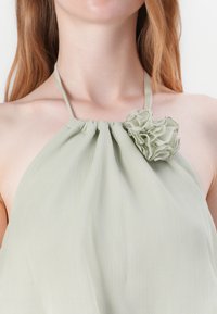 Svijetlozelena halter majica s nabranim dekolteom i teksturalnim cvjetnim detaljem, izrađena od lepršavog materijala s suptilnim sjajem.