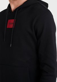 Sudadera con capucha negra que presenta un parche de logo rectangular rojo, cordones ajustables y un bolsillo frontal tipo canguro. Textura de tela suave.