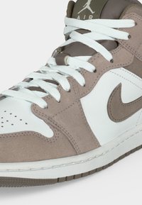 Καφέ και λευκό παπούτσι Air Jordan με λευκά κορδόνια, το λογότυπο Nike και διάτρητη περιοχή στη μύτη, σε απλό φόντο.