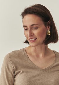 Pull beige à encolure en V avec une texture lisse. Une boucle d'oreille triangulaire dorée à l'oreille droite ajoute une touche audacieuse. Les cheveux sont coiffés en un carré soigné.