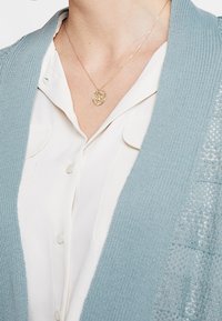 Ljusblå kofta med öppen stickad detalj över en vit skjorta med knappar. Guldsmycke med ett runt hängsmycke vilar vid halsen.