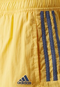Gula träningsshorts med elastiskt midjeband, med tre blå vertikala ränder och en Adidas-logotyp på nedre vänstra sidan. Strukturerad tyg.