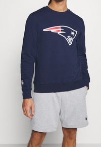 Sweatshirt marine avec un grand logo blanc et rouge des New England Patriots. Présente un col rond et des poignets côtelés. Porté avec un short gris.