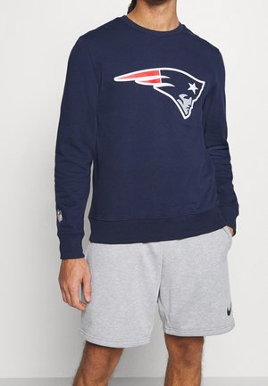 Sweatshirt marine avec un grand logo blanc et rouge des New England Patriots. Présente un col rond et des poignets côtelés. Porté avec un short gris.