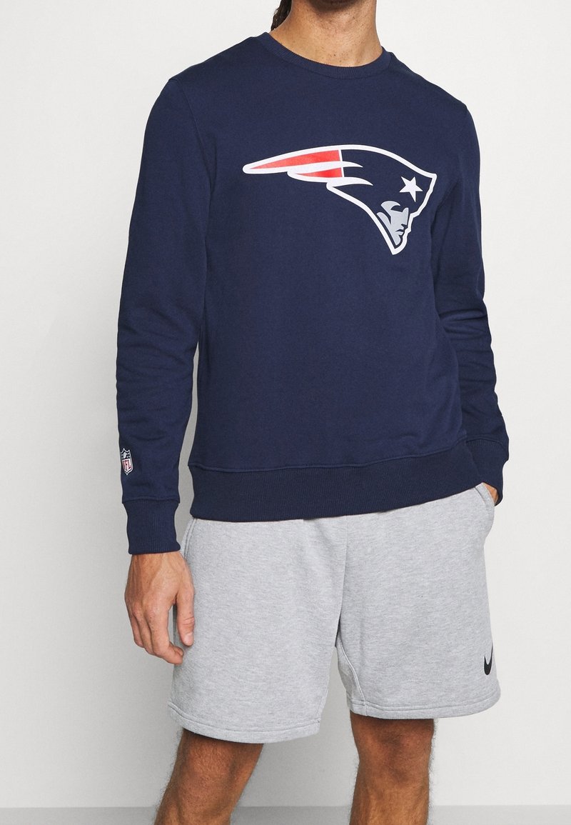 Sweatshirt marine avec un grand logo blanc et rouge des New England Patriots. Présente un col rond et des poignets côtelés. Porté avec un short gris.