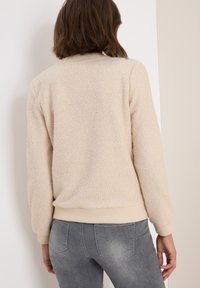 Frau in beiger, strukturierter Fleecejacke und ausgebleichten grauen Jeans, die mit dem Rücken zur Kamera vor einer weißen Wand steht.