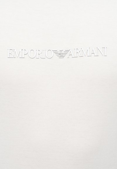 Λευκό ύφασμα με ανάγλυφη λευκή επιγραφή "EMPORIO ARMANI" και το λογότυπο του αετού της μάρκας ανάμεσα στις λέξεις.