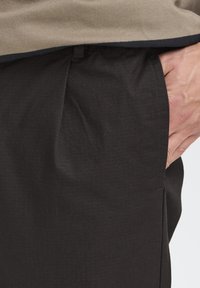 Mustat housut, joissa on hienovarainen teksturoitu kuvio ja sivutasku. Beige toppi peittää osittain vyötärönauhan.