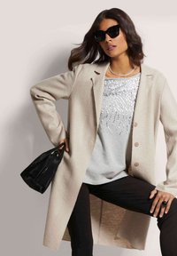 Beiger, strukturierter langer Mantel mit Knöpfen über einem grauen Pailletten-Oberteil; kombiniert mit schwarzen Hosen und einer kleinen schwarzen Handtasche; Sonnenbrille als Accessoire.