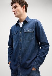 Tums ar pogām izgatavots tumši zils džinsu krekls, kas satur divas krūšu kabatas, izgāztu apkakli un garas piedurknes ar pogu aprocēm. Gluda tekstūra.