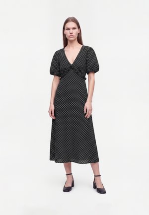 Vrouw draagt een zwarte midi-jurk met witte polkadotjes, pofmouwen, diepe V-hals en zwarte enkelsluiter-hakken, staand op een witte achtergrond.