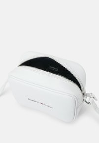 Bolso bandolera sintético blanco con cierre de cremallera, correa única y logo en relieve de "Tommy Jeans" en la parte delantera. Textura suave y diseño redondeado.