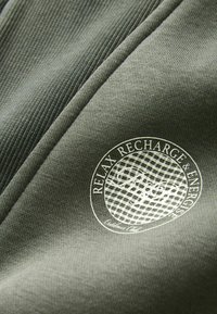 Tissu vert foncé avec une texture lisse, affichant un logo rond portant la mention "RELAXEZ, RECHARGEZ & ÉNERGISEZ" en impression blanche.