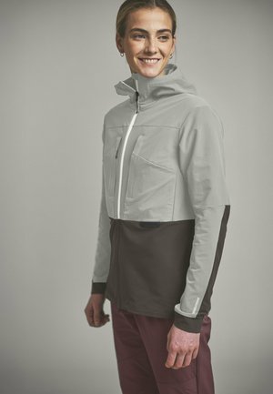 Junge Person lächelt, trägt eine hellgraue und dunkelbraune, langärmelige Zip-up-Jacke und weinrote Hose, steht vor einem einfarbigen Hintergrund.