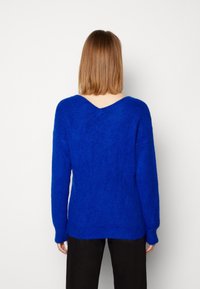 Pull tricoté bleu avec encolure en V, manches longues et finition texturée. Le dos présente une découpe en V profonde, l'ourlet est légèrement plus long à l'arrière.