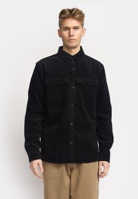 Zwart corduroy overhemd met een kraag, twee borstzakken, knopen aan de voorkant en een ribbeltextuur. Draag het over beige broek.