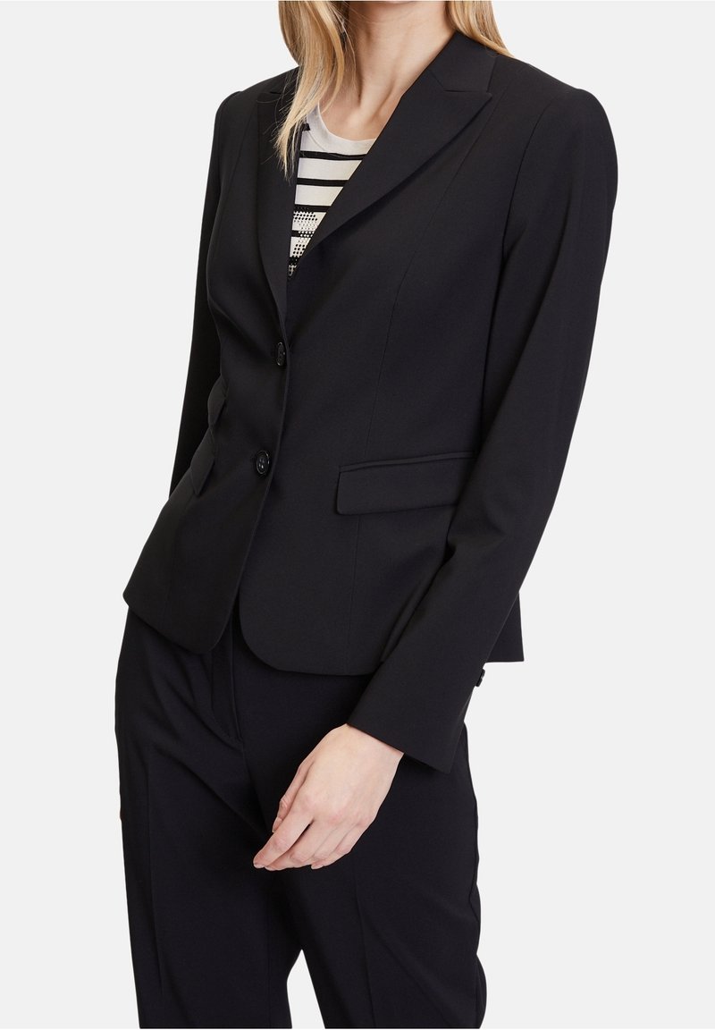 Femme portant un blazer noir ajusté à deux boutons sur un haut rayé noir et blanc, associé à un pantalon noir assorti.