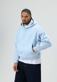 Sweat à capuche bleu clair en matière douce, avec une grande poche avant, des manches côtelées et une capuche à cordon. Porté sur une chemise blanche.