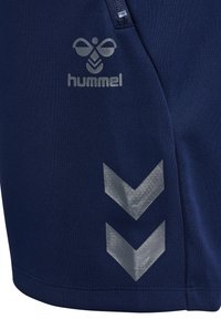 Marineblaue Sportshorts mit Hummel-Logo, versehen mit silbernen Chevron-Mustern und einer glatten, leichten Stoffstruktur. Mit Reißverschlusstasche.