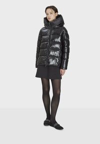 Svart puffjacka med blank finish, luva och dragkedja, med vadderade sektioner och oversized passform. Bärs över en svart kjol.