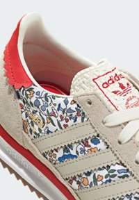 Adidas-sneaker med en beige och blommönstrad textilöverkonstruktion, röda accenter och vågiga vita ränder. Har en texturerad tunga och snörning.
