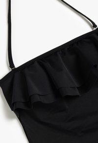 Haut de bikini noir avec une bande volantée, des bretelles fines ajustables et une texture lisse. Le tissu semble extensible et ajusté.