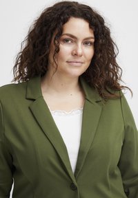 Oliwkowa marynarka z wycięciem, pojedynczym czarnym guzikiem i gładką teksturą. Noszona na białej bluzce z koronkowym wykończeniem.