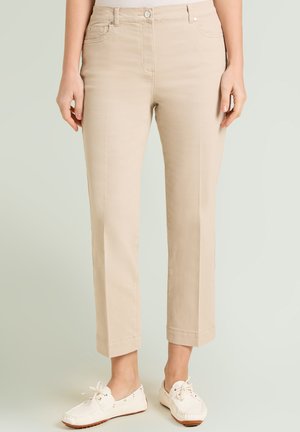 Vrouw die beige rechte broek en witte casual veterschoenen draagt, staand tegen een effen lichte achtergrond.