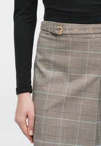 Brun ruter skjørt med gullfarget beslag, et belte med spenne og lyseblå detaljer, kombinert med en svart ribbestrikket langermet topp.