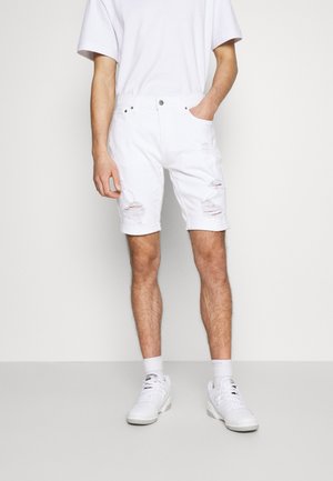 Homme portant un short en denim blanc usé, des baskets blanches, des chaussettes blanches et un T-shirt blanc, debout devant un fond clair uni.