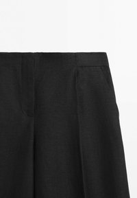 Primo piano di pantaloni neri a vita alta che mostrano la cintura, la piega frontale e la tasca laterale su uno sfondo bianco.