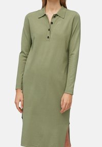 Robe polo à manches longues de couleur vert olive en tissu doux, avec un col à boutons, trois boutons noirs, et des fentes latérales pour faciliter le mouvement.
