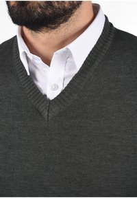 Pull en maille gris foncé avec col en V, présentant des détails côtelés autour du col, superposé à une chemise blanche à boutons.