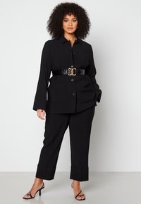 Zwart op maat gemaakte jumpsuit met knoopsluiting aan de voorkant, grote zakken en een taille die is aangetrokken met een glanzende zwarte riem met gouden accenten.