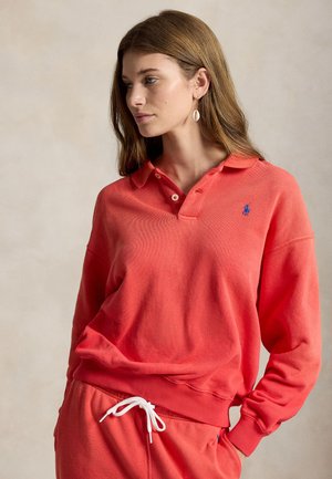 Nő hosszú barna hajjal, korallszínű póló pulóvert és hozzáillő szabadidőnadrágot visel, fehér húzózsínnel, semleges háttér előtt.