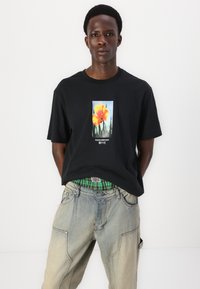 Sort bomulds T-shirt med et frontgrafik af gule blomster. Kombineret med slidte, blå baggy-jeans med et ternet taljebånd.