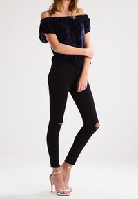 Niebieska, welurowa bluzka z odkrytymi ramionami z teksturowanym wzorem, w połączeniu z czarnymi, skinny jeansami z przetarciami i srebrnymi, rzemykowymi szpilkami.