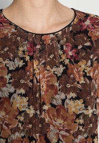 Kvinna i en blommig blus med veck, med stora bruna, gula och röda blommor på en svart bakgrund.