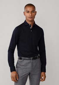 Mornarsko plava majica s dugim rukavima i gumbima na ovratniku, uparena s sivim teksturiranim hlačama i crnim remenom sa srebrnom kopčom.