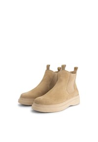 Een paar tan suède Chelsea boots met trektabs en dikke beige rubber zolen op een witte achtergrond.
