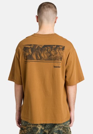 Mand med kort hår iført en brun Timberland T-shirt med et grafik af flere støvler på ryggen, camouflagebukser synlige nedenunder.