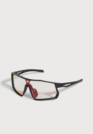 Lunettes de sport noires avec de grands verres transparents, un pont de nez rouge et des branches ventilées, sur fond clair.