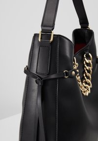 Sac à main en cuir noir avec détail chaîne dorée, sangle noire, et accent décoratif en cuir noué sur fond gris uni.