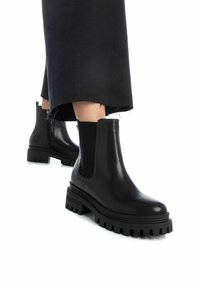 Botas de cuero negras hasta el tobillo con paneles laterales elásticos y suelas de goma gruesas, con una superficie lisa y acentos texturizados en la suela.