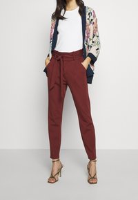 Frau trägt bordeauxfarbene hoch taillierte Papiertütenhose, weißes ärmelloses Top, florale Kimono-Jacke und olivgrüne Sandalen mit Absatz und Riemchen.