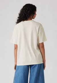 Beiges Oversize-T-Shirt mit kurzen Ärmeln, aus weichem Stoff gefertigt. Es verfügt über einen klassischen Rundhalsausschnitt und einen geraden Saum, kombiniert mit hellblauen Jeans.