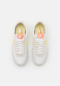 Bílé a žluté tenisky Nike s texturovanou béžovou platformou, bílými tkaničkami a kontrastním oranžovým logem na jazyku.