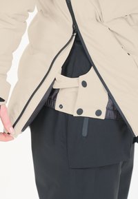 Beige vadderad jacka med dragkedja, lagd över svarta byxor. Har tryckknappsdetaljer och elastiska detaljer vid fåll och ärmslut.