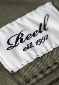 Etiqueta blanca con texto en cursiva negra "Reell" y "est. 1997" sobre un fondo de tela verde oliva texturizado, con bordes cosidos.