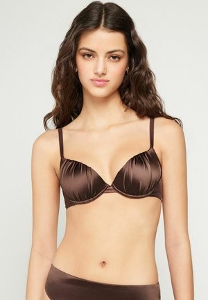 Junge Frau mit langen, welligen Haaren, die einen braunen Satin-BH und passende Unterteile trägt, blickt auf einen schlichten Hintergrund.